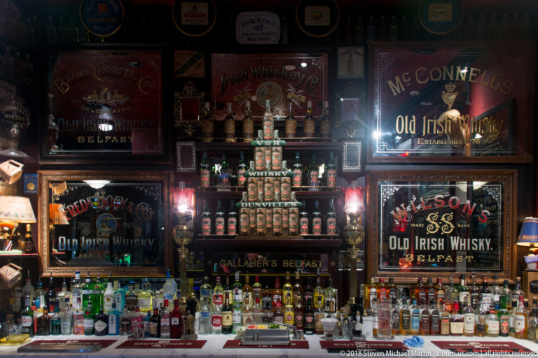 Belfast bar crowded with bottles, mirrors, and historic Old Irish Whisky advertising panels under warm lamps.