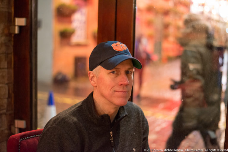 Seated man in a dark sweater and baseball cap inside a warmly lit Belfast pub.