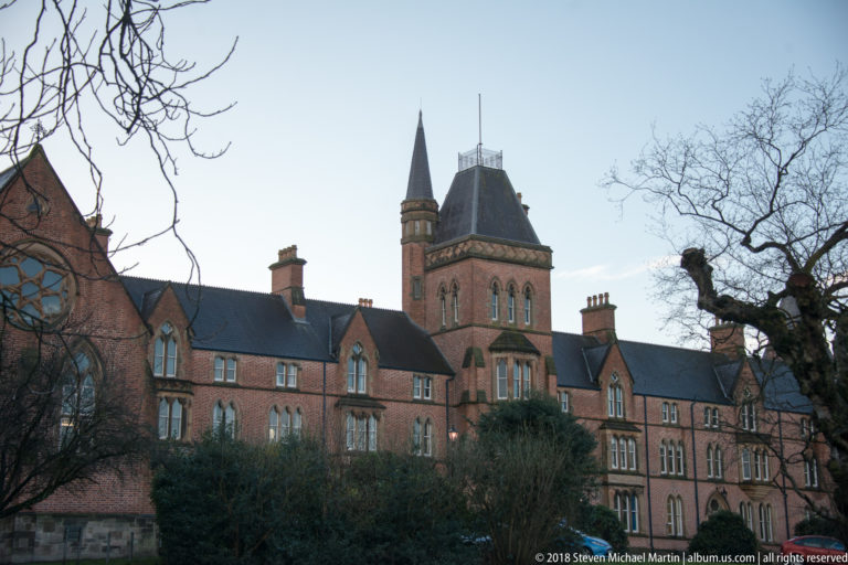 Long red-brick collegiate-style building with gables, chimneys, and a slender spire behind winter trees.