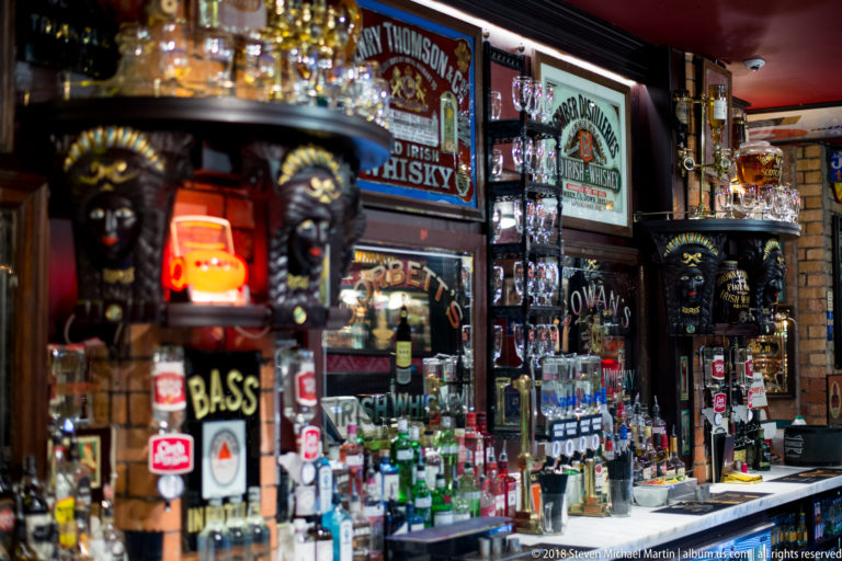 Belfast pub bar filled with bottles, taps, mirrors, vintage drink signs, and carved decorative figures.
