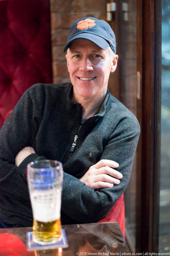 Smiling man in a baseball cap seated in a Belfast pub with a partly filled pint glass in front.