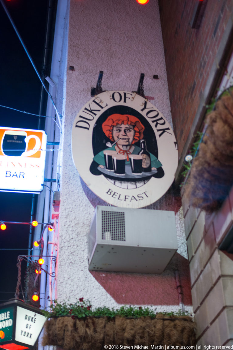 Round Duke of York Belfast pub sign mounted high on a white wall above a decorated alley.