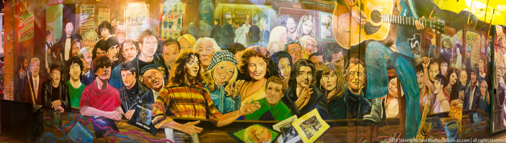 Large colorful mural of many faces and figures extending along a narrow Belfast passage wall.