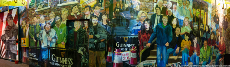 Long Belfast mural with painted people, Guinness signs, and a pub entrance motif along a narrow passage.