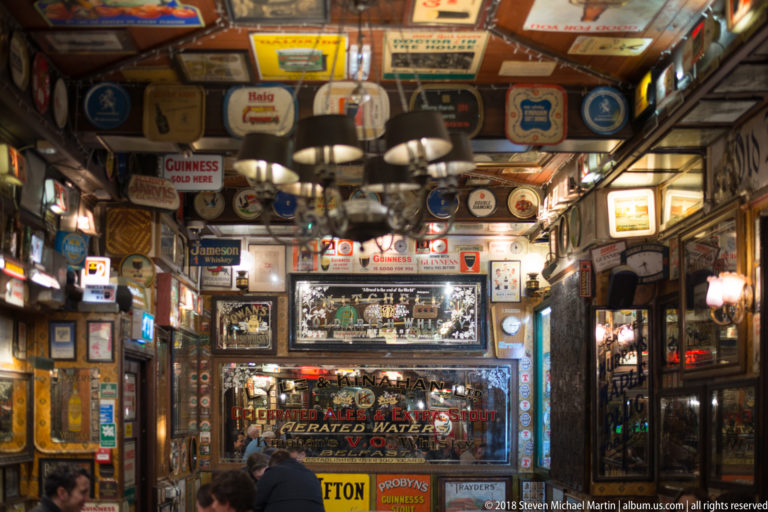 Belfast pub interior densely covered with vintage signs, mirrors, enamel advertisements, and hanging memorabilia above seated patrons.