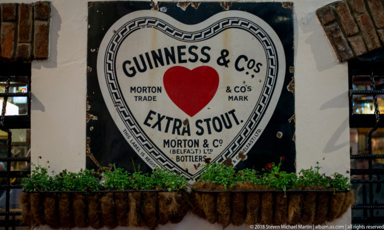 Heart-shaped Guinness & Co’s Extra Stout wall advertisement with Morton & Co Belfast bottlers lettering and flower boxes below.