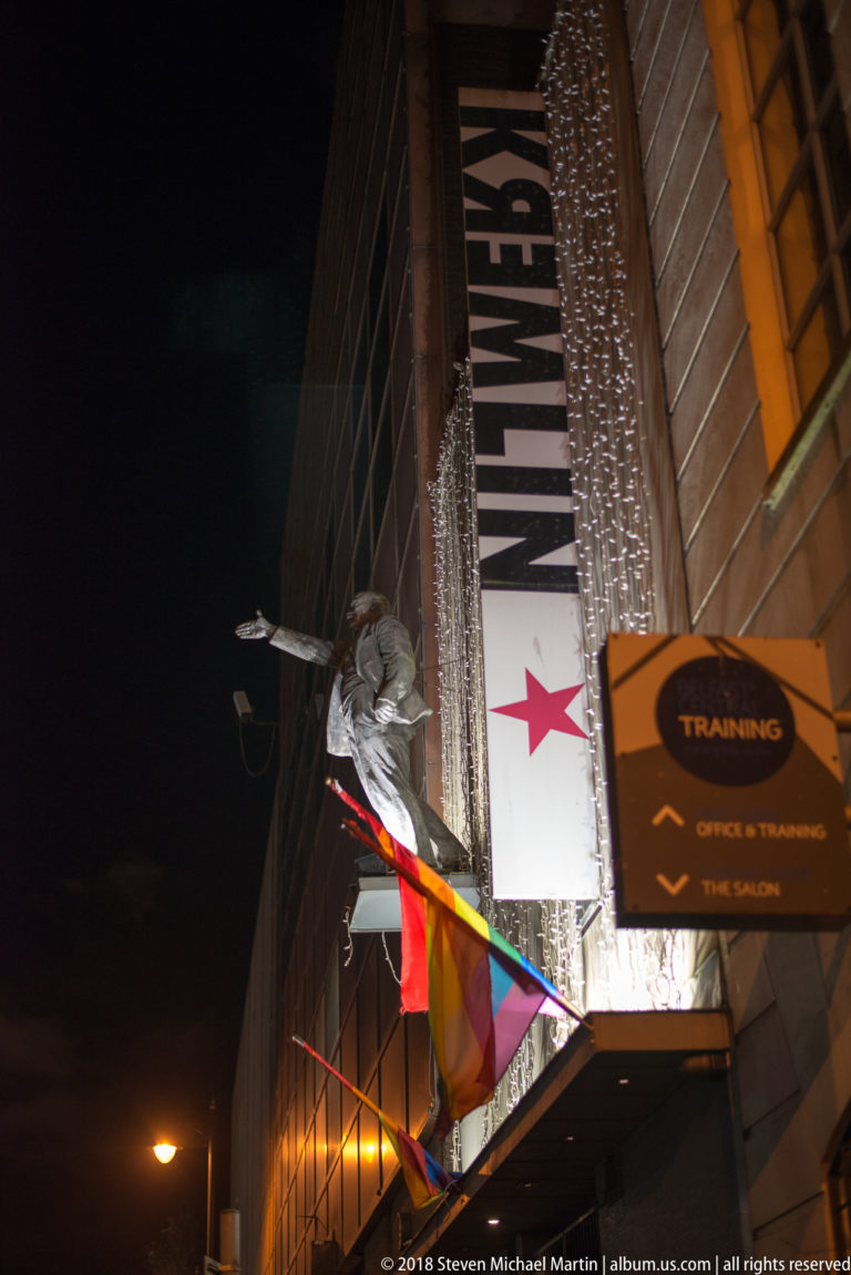Nighttime Belfast venue entrance with vertical sign, metallic fringe, rainbow fabric, and a suspended sculptural figure.