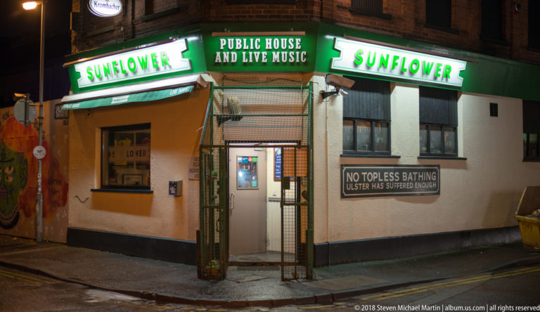 Sunflower pub corner at night with green Public House and Live Music signs and humorous wall plaque.