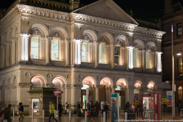 Illuminated historic stone civic building with columns, arched windows, pediment, and pedestrians at street level in Belfast.