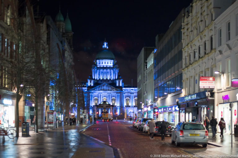 Wet Belfast street at night leading toward illuminated Belfast City Hall, with parked cars, shops, and pedestrians.