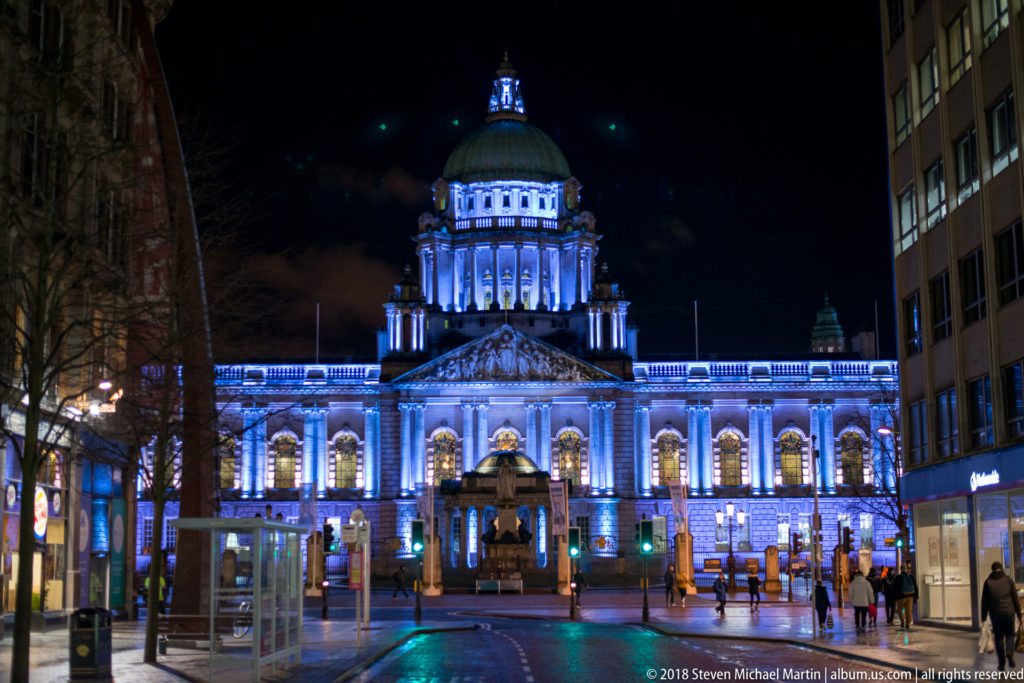 Belfast City Hall illuminated blue at night, with domed central block, symmetrical wings, wet pavement, and pedestrians.