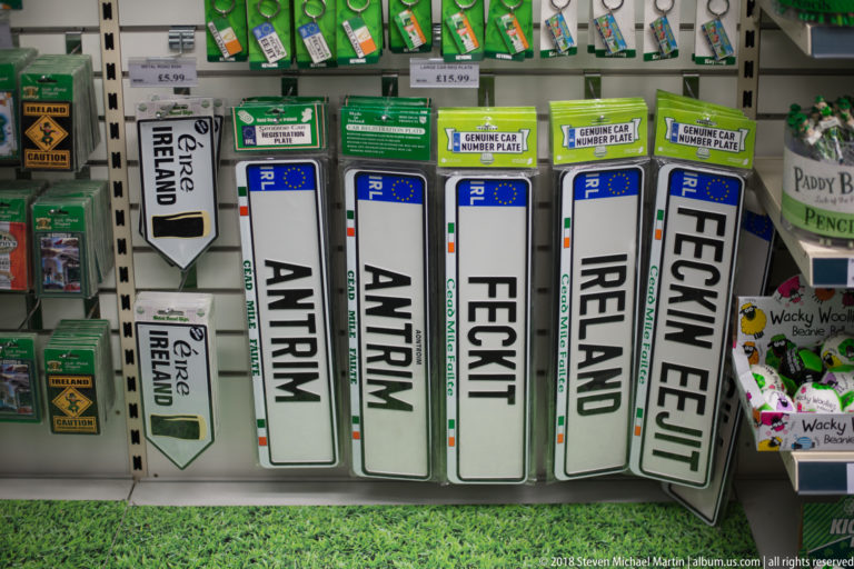 White novelty road-sign plaques reading Antrim, Feckit, Ireland, and Feckin Eejit on a Belfast souvenir-shop wall.