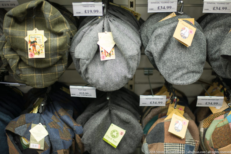 Rows of gray, brown, and checked tweed-style flat caps hanging on a Belfast shop wall with price tags.
