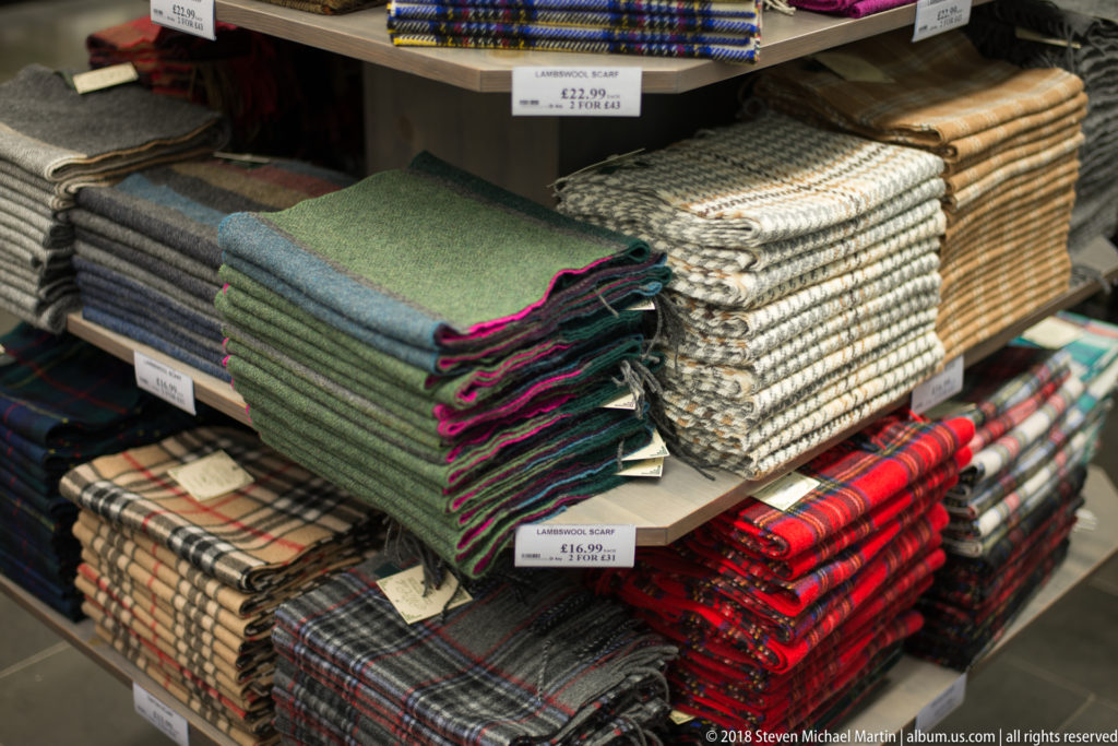 Stacks of green, cream, gray, and red tartan wool textiles folded on shelves in a Belfast shop.