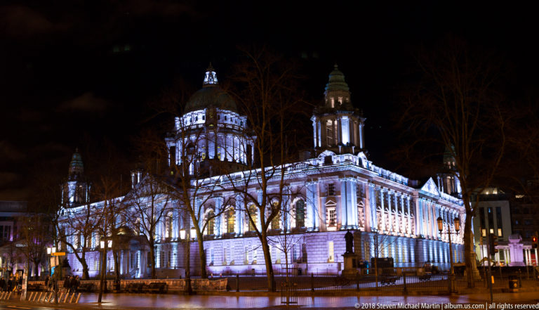 Oblique night view of Belfast City Hall with illuminated stone façades, corner towers, dome, and bare trees in the foreground.