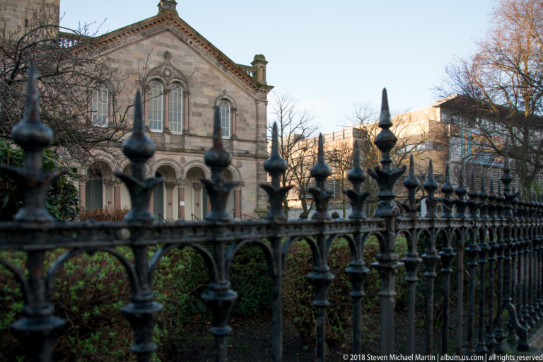 Ornate black iron railings in sharp foreground with a pale stone church façade and arched windows behind shrubs.