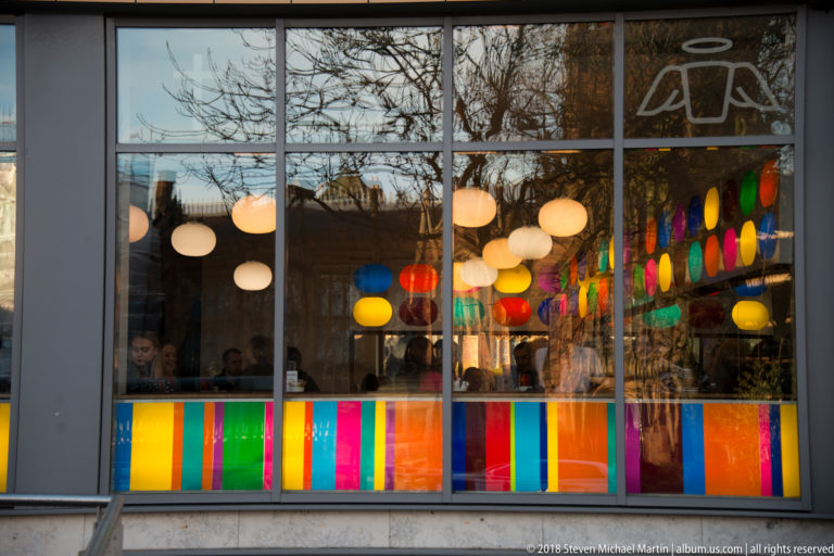 Large storefront window with colorful rectangular panels, round hanging lights, and reflections of trees and nearby buildings.