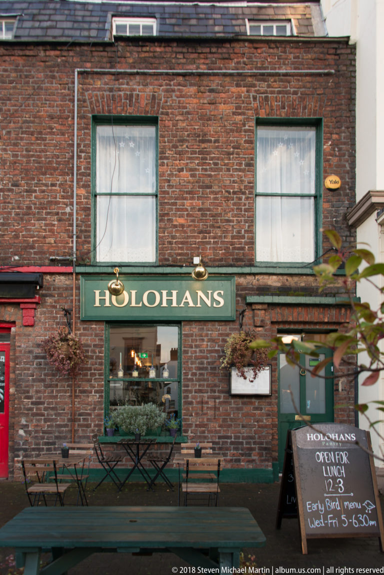 Brick exterior of Holohans with green sign, open service window, outdoor tables, and a chalkboard advertising lunch.