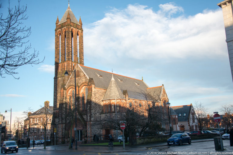 Large stone Gothic Revival church with tall square tower, long nave, and pointed windows at a Belfast street corner.