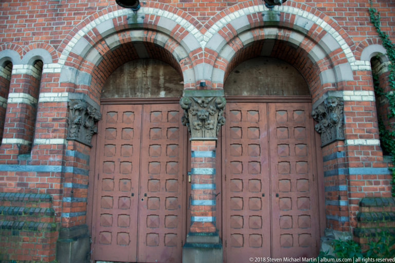 Two wooden doors beneath striped Romanesque arches with dark columns, carved capitals, and patterned red brickwork.