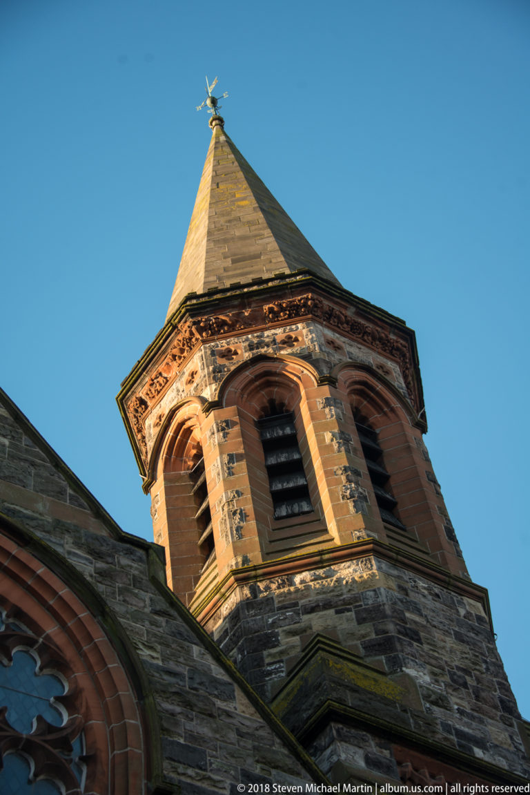 Low-angle close view of an octagonal stone church belfry with louvered arches, pyramidal roof, and weather vane.
