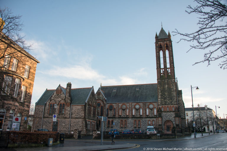 Large Belfast church in contrasting stone, with pointed windows, steep gables, and a tall open belfry tower beside the street.