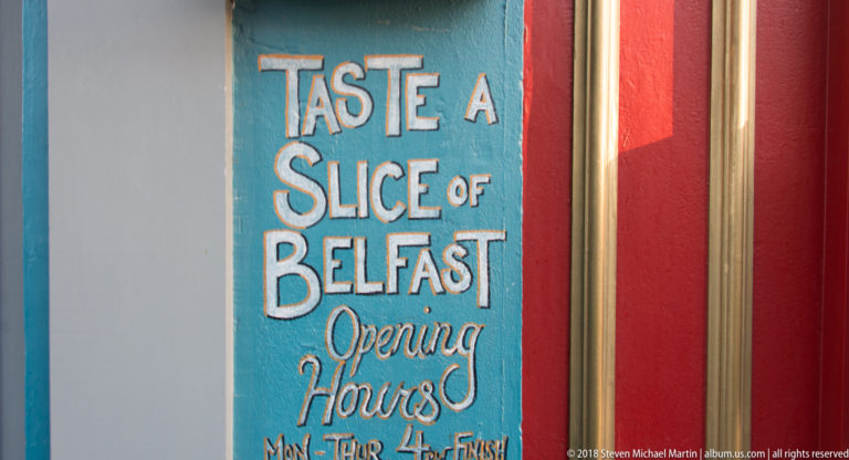 Hand-painted turquoise sign reading “Taste a Slice of Belfast” with opening hours, bordered by white and red panels.
