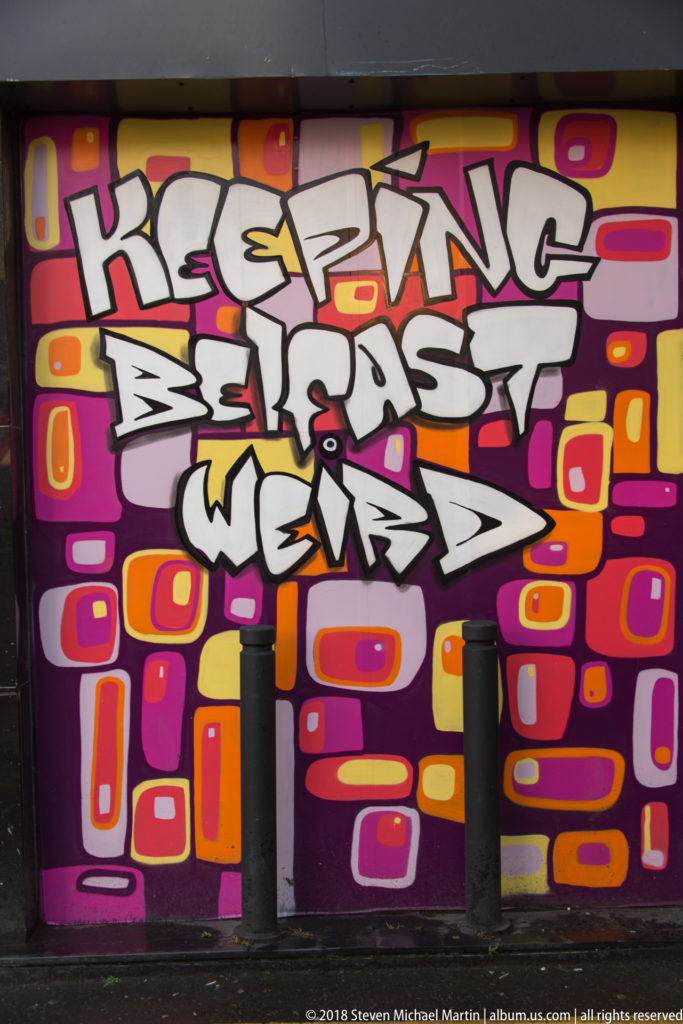 Colorful graffiti mural reading “KEEPIN BELFAST WEIRD” in white and black letters over pink, orange, red, and yellow shapes.
