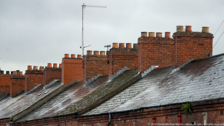 Rows of red-brick chimney stacks and chimney pots above weathered slate roofs on Belfast terraced houses.