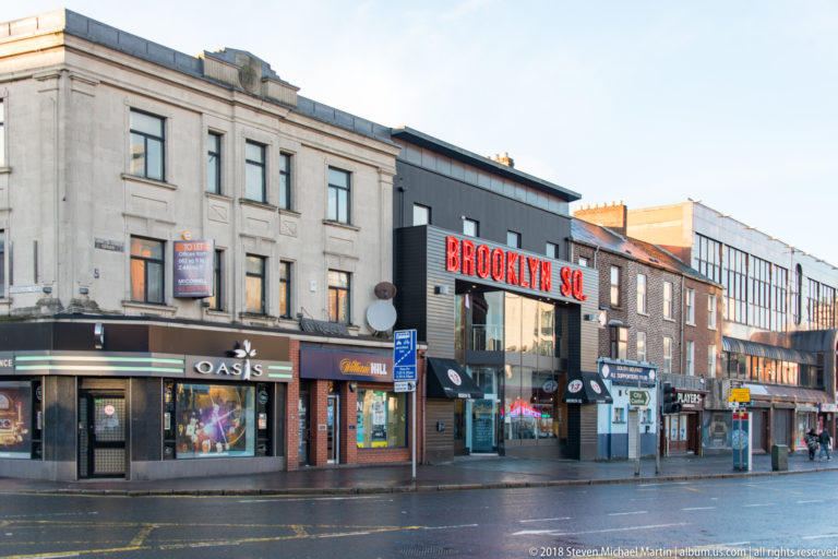 Belfast commercial street with the red “BROOKLYN SQ” sign above one storefront and neighboring Oasis and small businesses.
