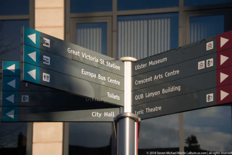 Black directional signpost lists Belfast destinations including City Hall, Ulster Museum, Great Victoria Street Station, and the Lyric Theatre.
