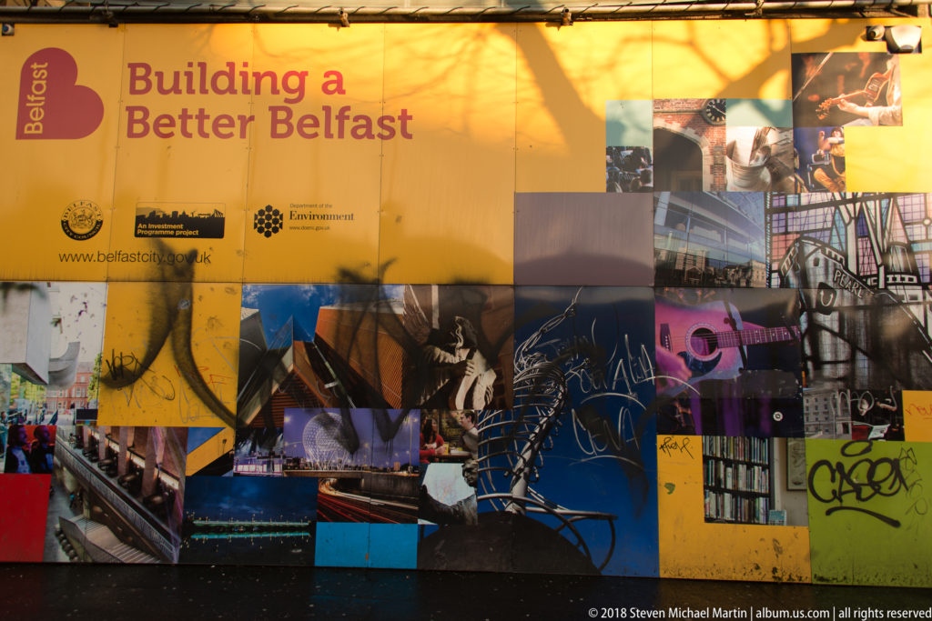 Yellow “Building a Better Belfast” construction hoarding covered with a collage of city photographs, logos, and graffiti.