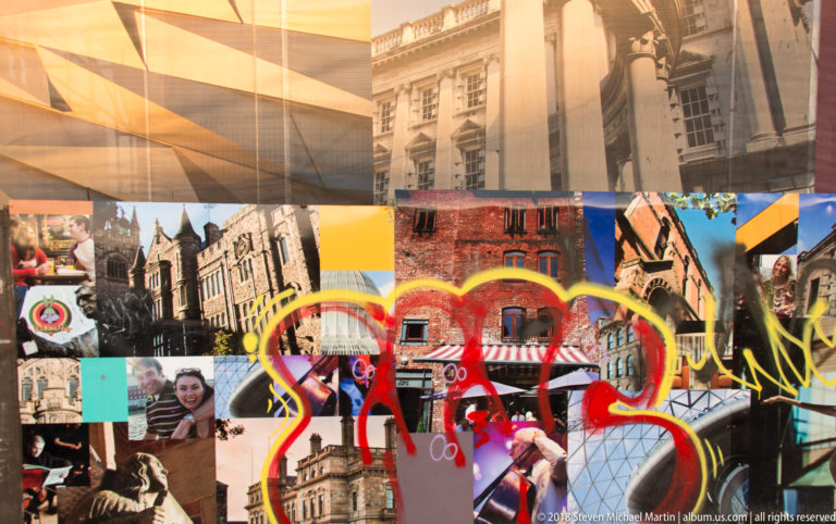 Close view of a Belfast redevelopment hoarding with architectural photographs, people, and bold red and yellow painted loops.