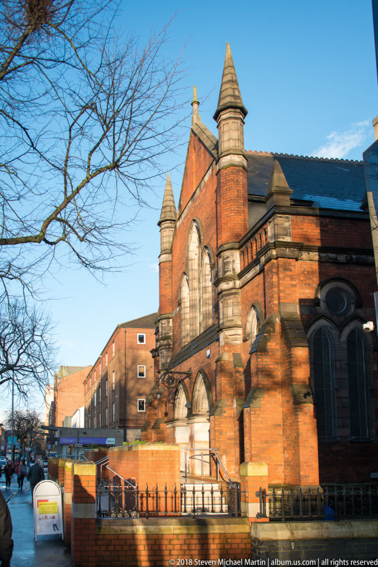 Red-brick Gothic Revival church with pointed windows, stone trim, and small pinnacles beside a Belfast street.