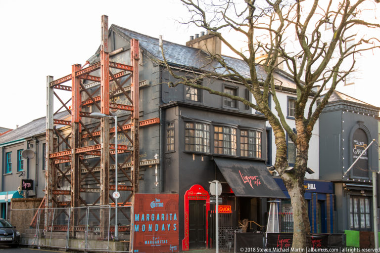 Dark-painted Belfast pub frontage with red exterior metal stairs, Guinness branding, awnings, and a leafless street tree.