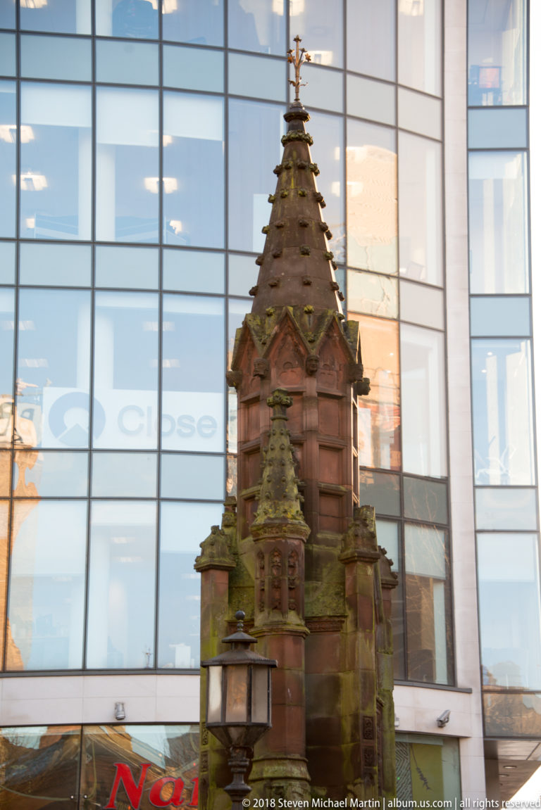 Ornate red-sandstone Gothic Revival street monument with a steep pointed cap standing before a modern glass building.