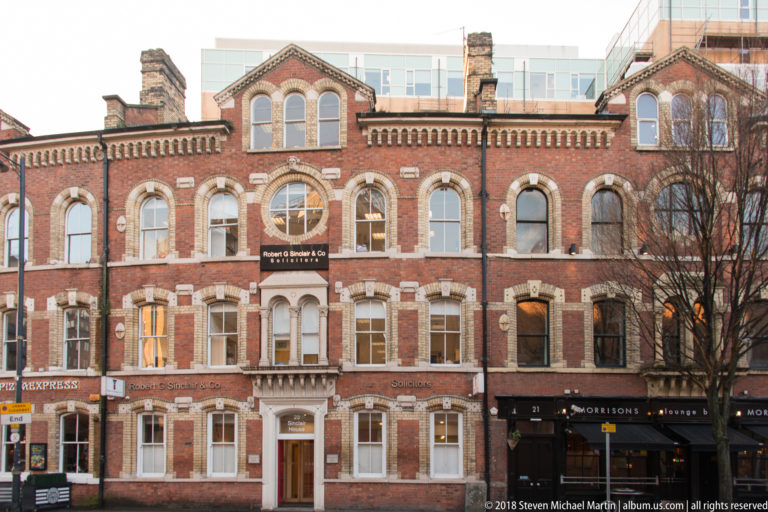 Broad red-brick historic commercial building with pale stone trim, arched windows, central balcony, and ground-floor shops.