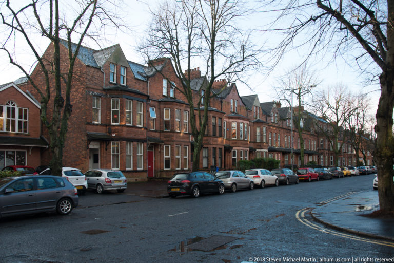 Red-brick Belfast houses with bay windows and gables line a wet street filled with parked cars.