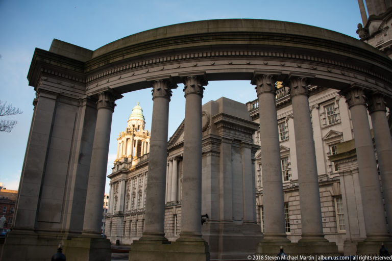 Belfast City Hall dome and tower framed between tall classical columns of a curved foreground colonnade.