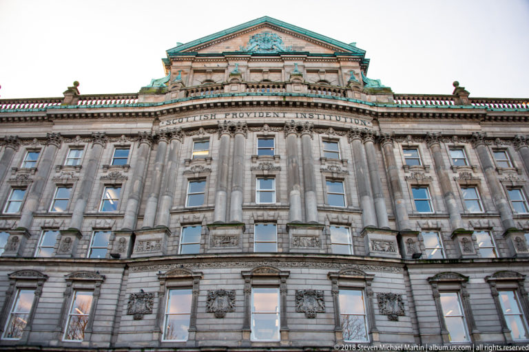 Upper facade of the Scottish Provident Institution with carved stonework, columns, pediment, and green-patinated copper details.