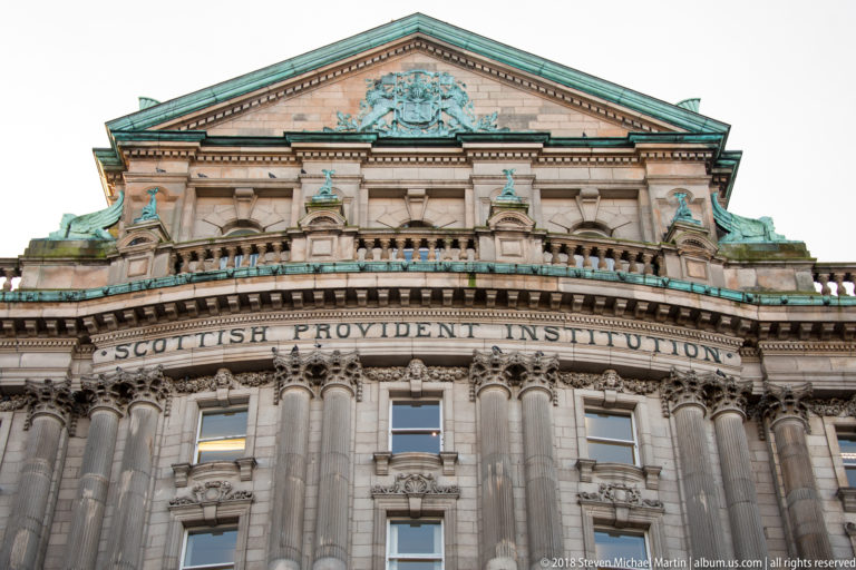 Close upward view of the Scottish Provident Institution pediment, carved name band, columns, balustrade, and green roof trim.