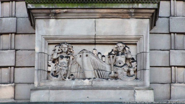 Rectangular stone relief with two carved figures flanking a draped central object, set into an ashlar facade.