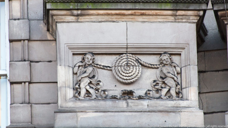 Stone facade relief with two carved figures holding or flanking a large spiral motif within a rectangular frame.