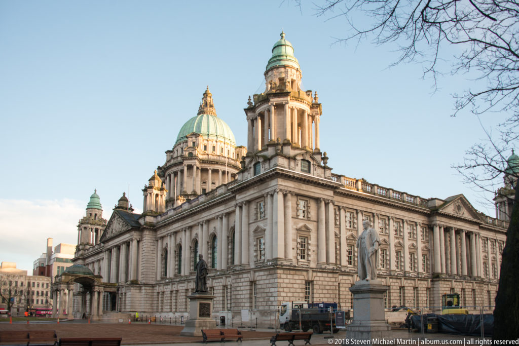 Three-quarter view of Belfast City Hall with pale stone facades, green copper dome, corner towers, statues, and open plaza.