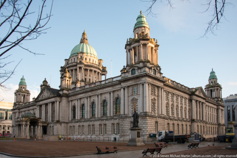 Angled view of Belfast City Hall with long stone facade, green dome and tower, foreground statue, benches, and vehicles.