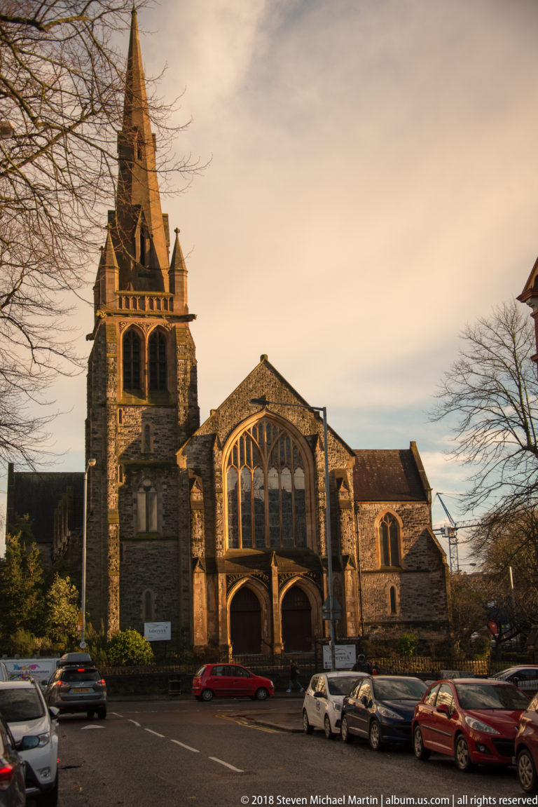 Stone Gothic Revival church with a square tower, large traceried window, and cars parked in front.