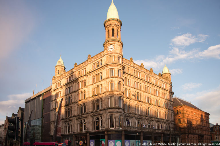 Ornate pale-stone corner building glowing in low sunlight, with rows of arched windows and a tall cylindrical turret.