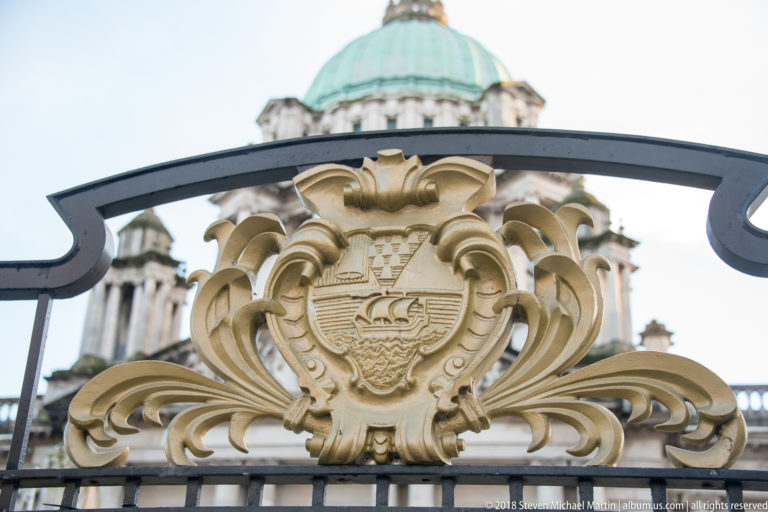 Gilded Belfast civic crest with ship motif mounted on black ironwork, with the City Hall dome blurred behind.