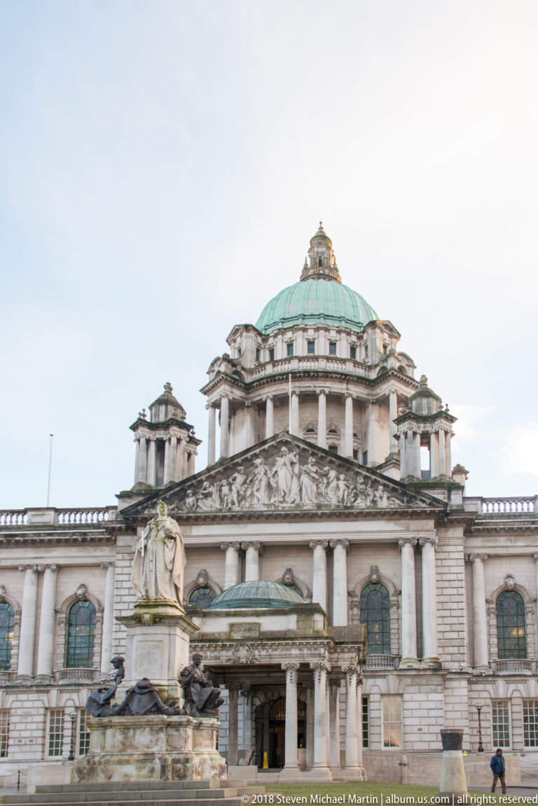Front view of Belfast City Hall entrance, sculpted pediment, green dome, and a large memorial statue in the foreground.