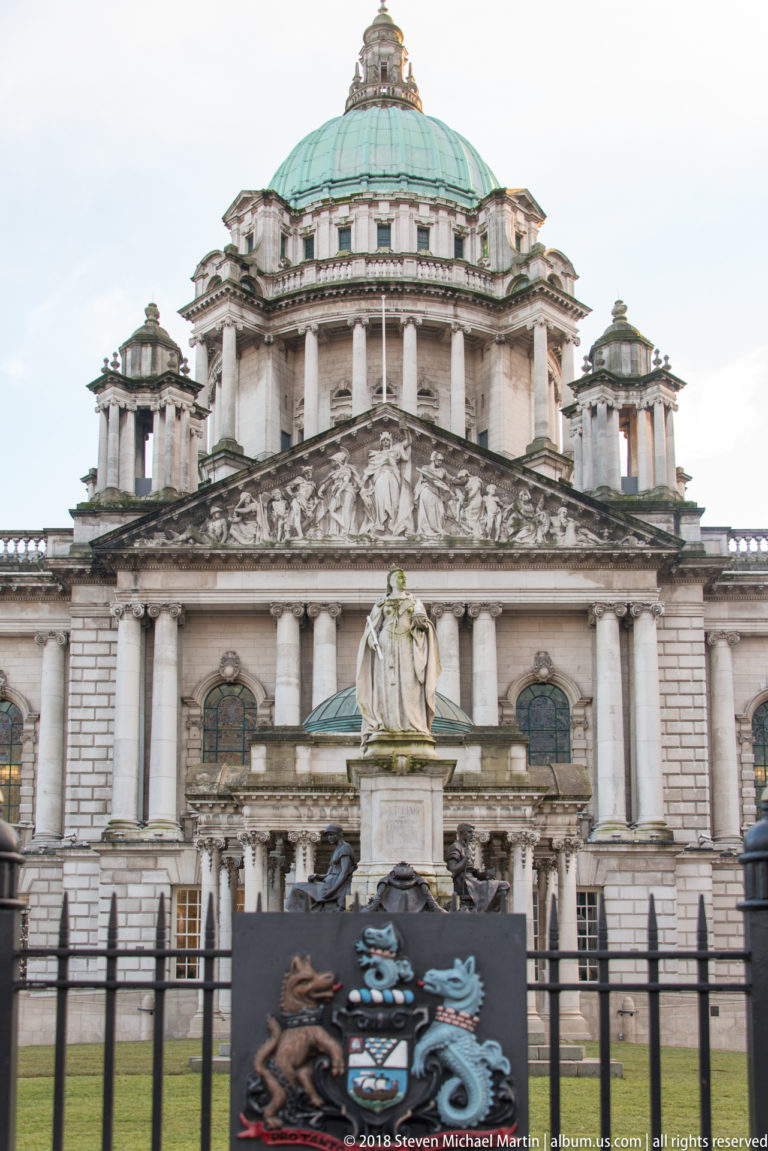 Belfast City Hall centered behind an iron fence and heraldic panel, with its green dome and pediment above.
